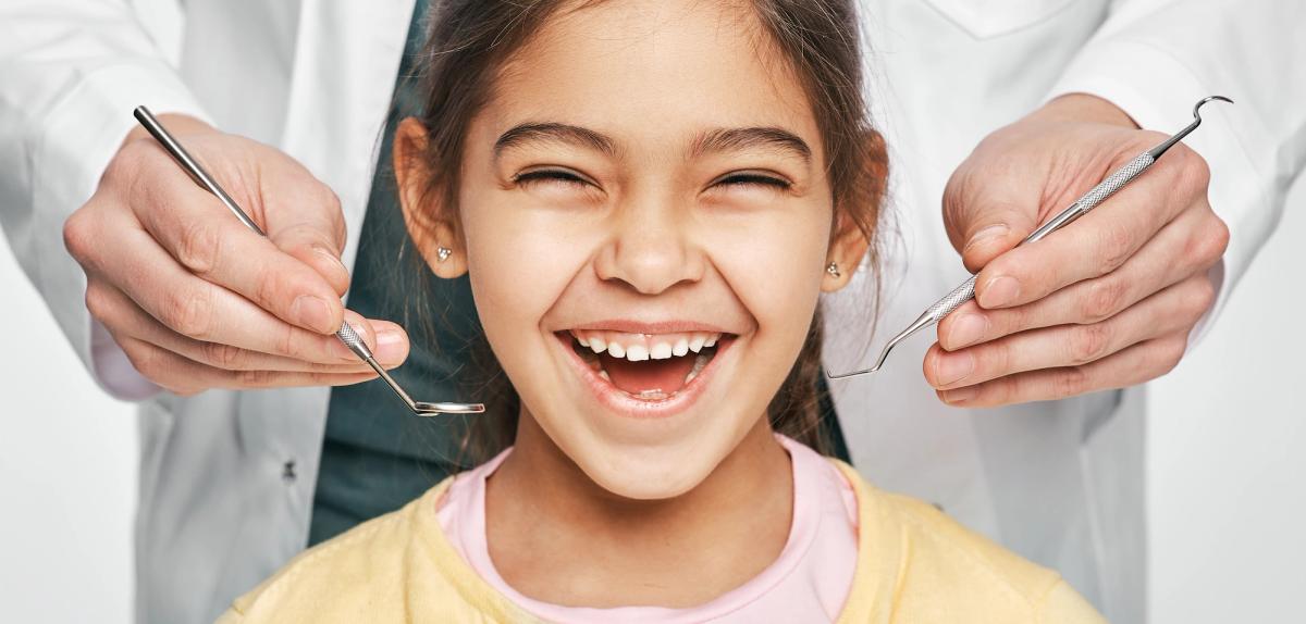 A happy young girl smiles widely as a dentist's hands hold dental tools.