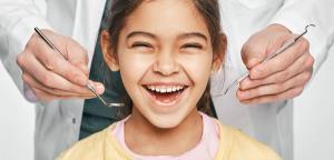 A happy young girl smiles widely as a dentist's hands hold dental tools.