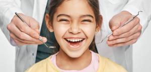 A happy young girl smiles widely as a dentist's hands hold dental tools.