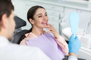 A happy woman in a dentist's chair smiles at her reflection in a mirror held by a dentist.