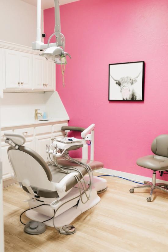 A modern dental office featuring a pink wall with a Highland cow art print blowing a pink bubble, a gray dental chair, and white cabinets.