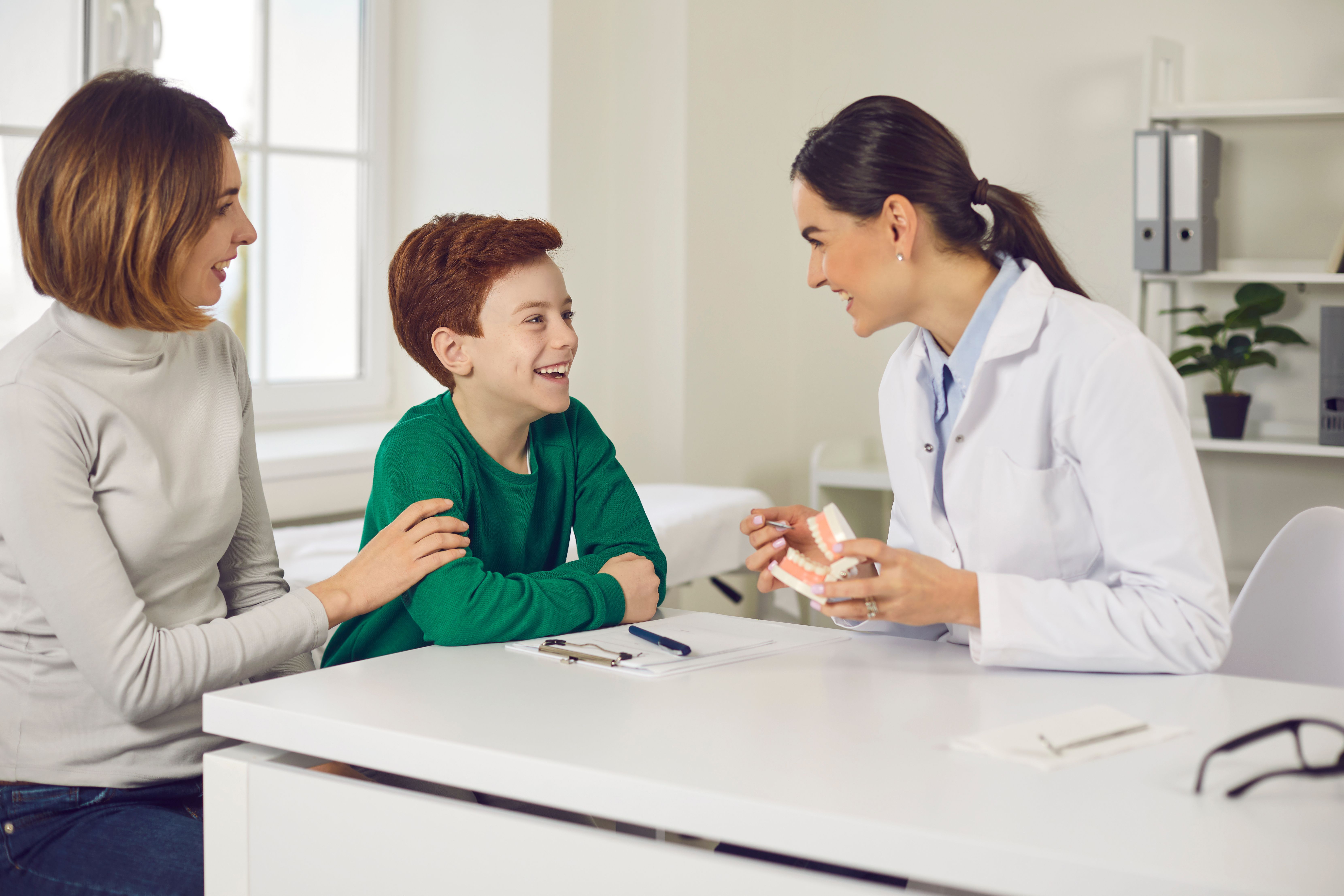 A female dentist explains a tooth model to a smiling boy and his mother.