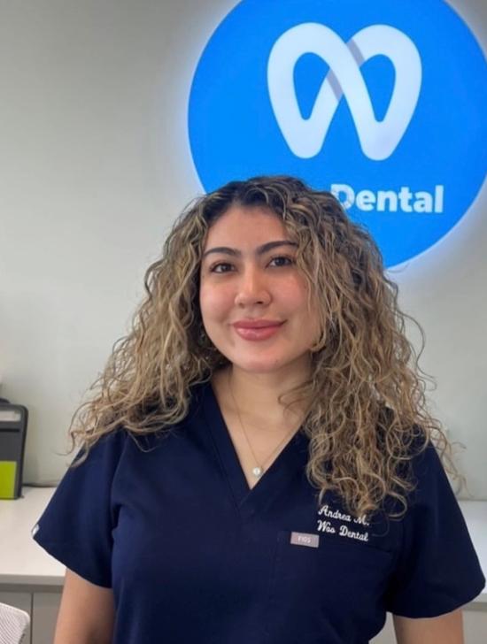 Andrea M. smiling in navy Woo Dental scrubs, with the Woo Dental logo behind her.