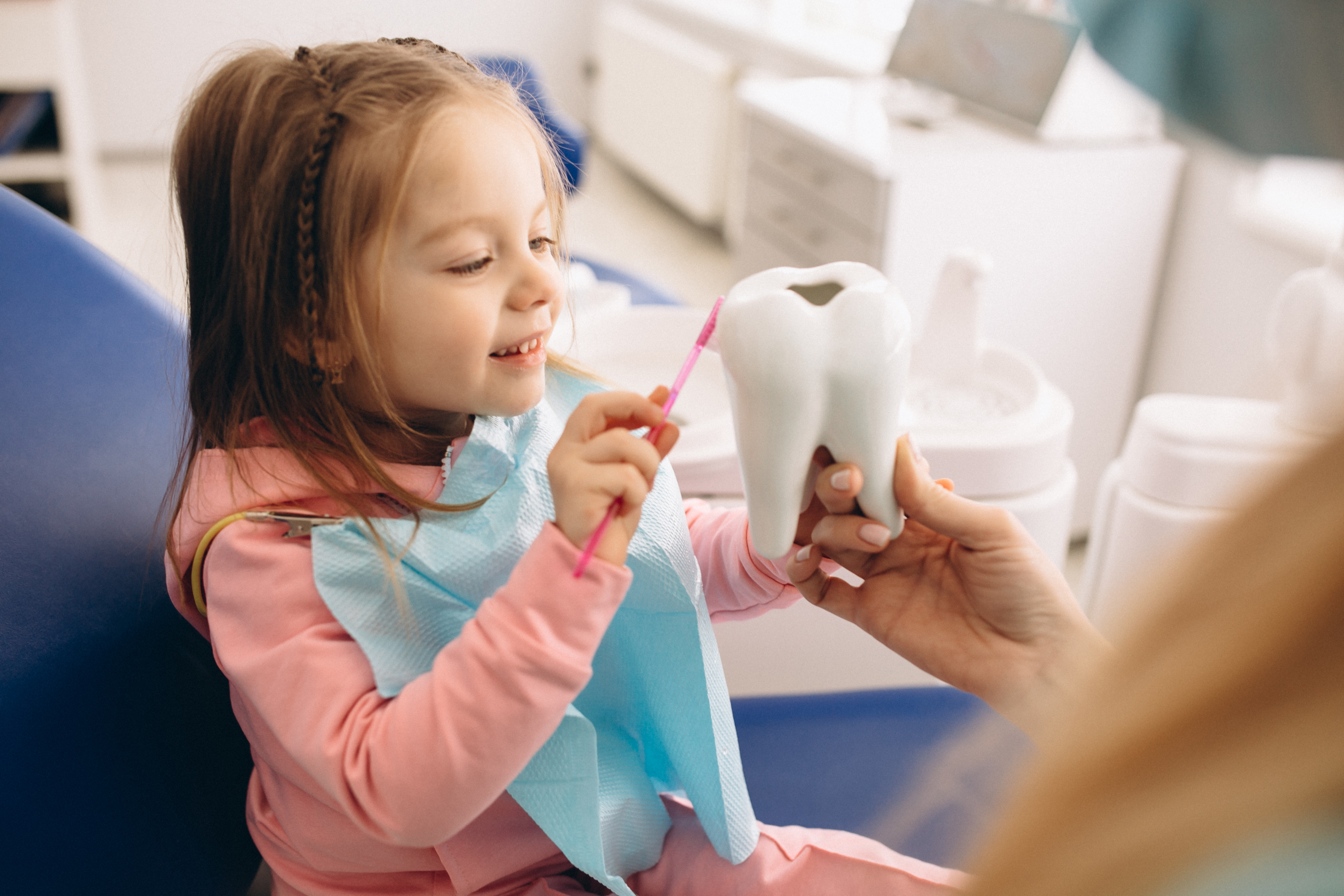 A happy young girl practices brushing a large model tooth with a pink toothbrush, guided by an adult.