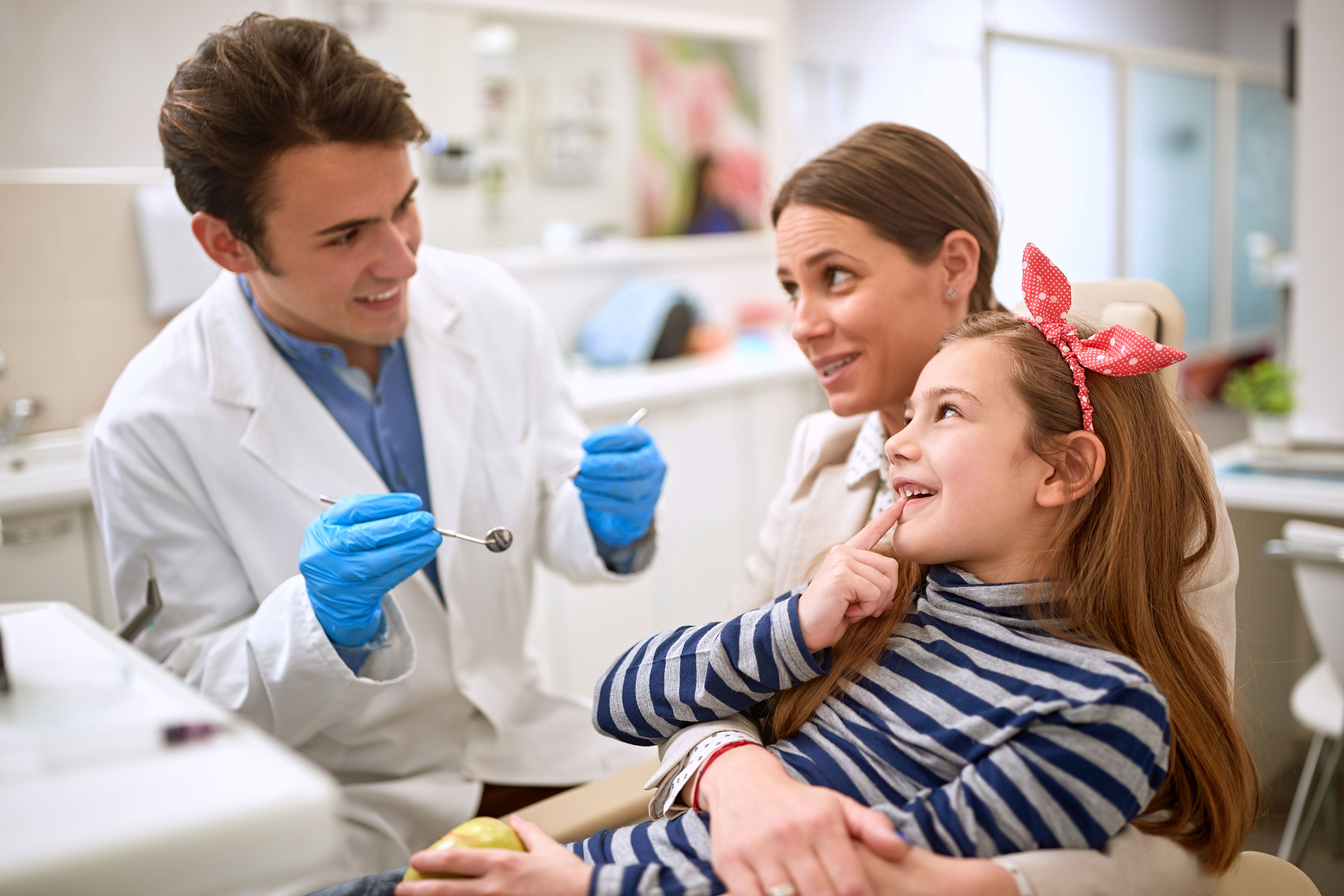A smiling dentist holds tools while looking at a young girl and her mother. The girl, sitting on her mother's lap, points to her tooth.