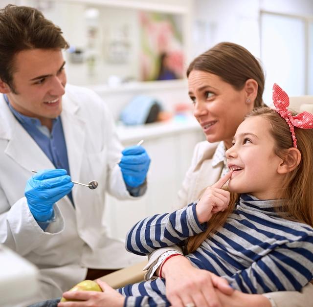 A smiling dentist holds tools while looking at a young girl and her mother. The girl, sitting on her mother's lap, points to her tooth.