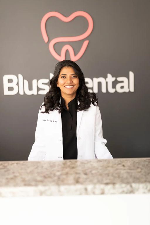 Smiling Dr. Sonia Prabhu, DDS, in a white lab coat, standing in front of the Blush Dental logo.