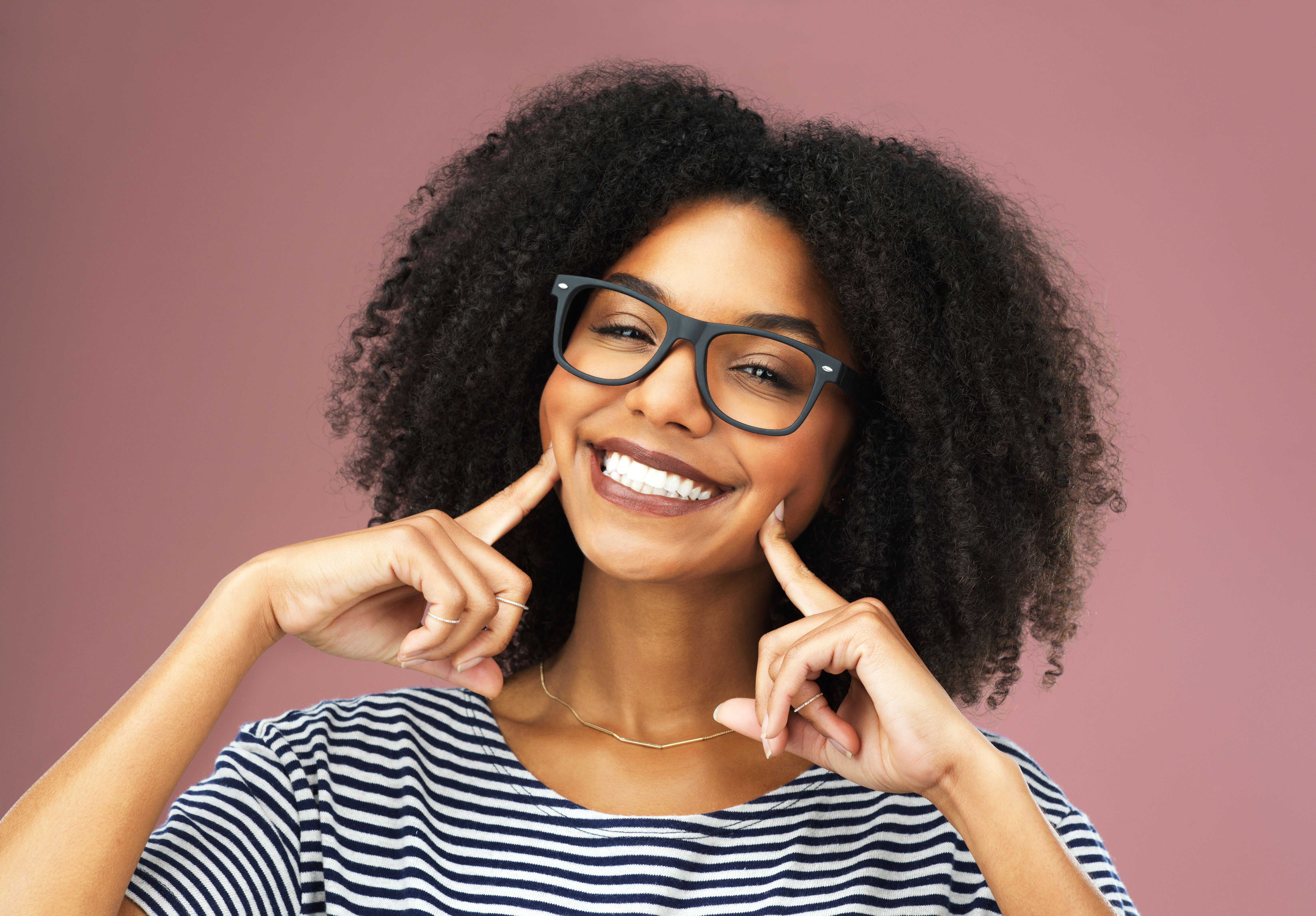 Black woman with curly hair and glasses smiling and pointing to her dimples.
