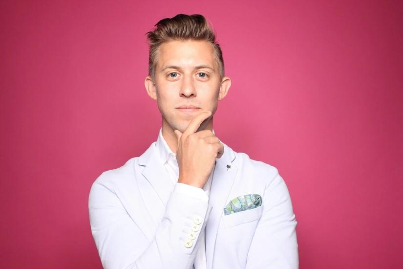 a man in a white suit is standing in front of a pink background with his hand on his chin .