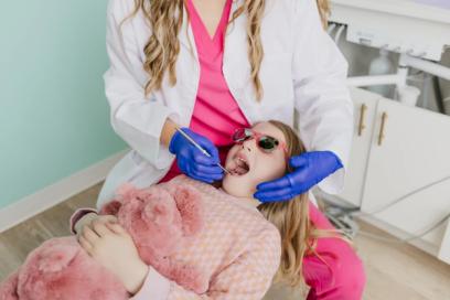 A dentist examines a young girl's teeth; she wears pink sunglasses and holds a pink stuffed animal.
