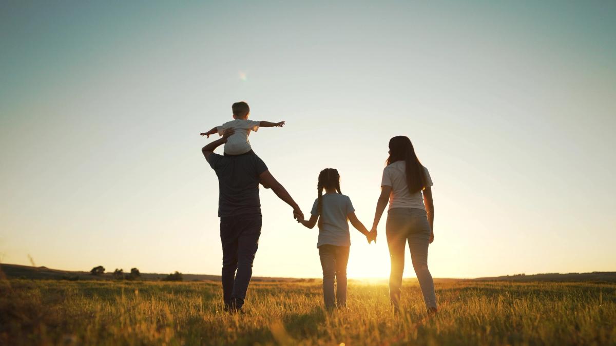 Silhouetted family of four walking through a grassy field at sunset, with a child on the father's shoulders.