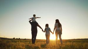 Silhouetted family of four walking through a grassy field at sunset, with a child on the father's shoulders.