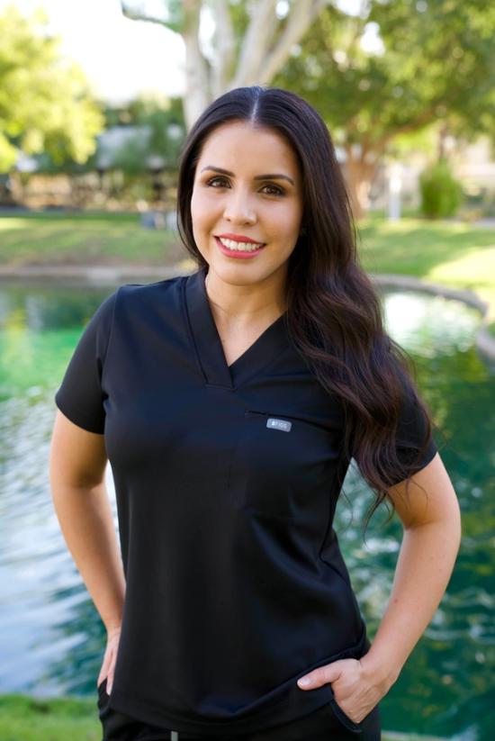 A smiling woman in black scrubs stands outdoors by a pond.