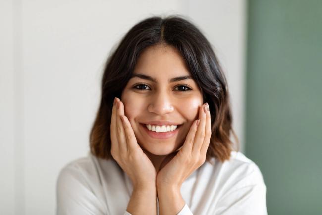A smiling woman with her hands on her cheeks looks happily at the camera.