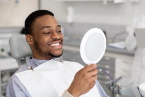 A smiling man in a dental chair looks at his reflection in a handheld mirror.