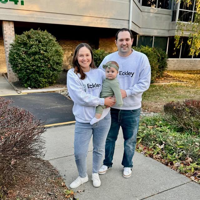 A smiling mother holding a baby, and a father, all wearing matching grey Eckley sweatshirts outdoors.