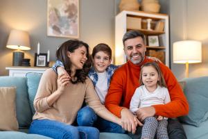 A smiling family of four sits together on a couch in their living room.