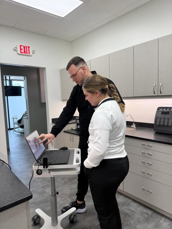 A man points at a dental 3D model on a laptop while a woman watches in a clinic.