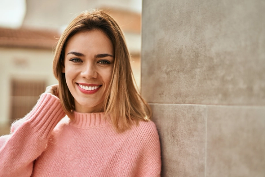 Smiling woman in a pink sweater leaning against a wall.