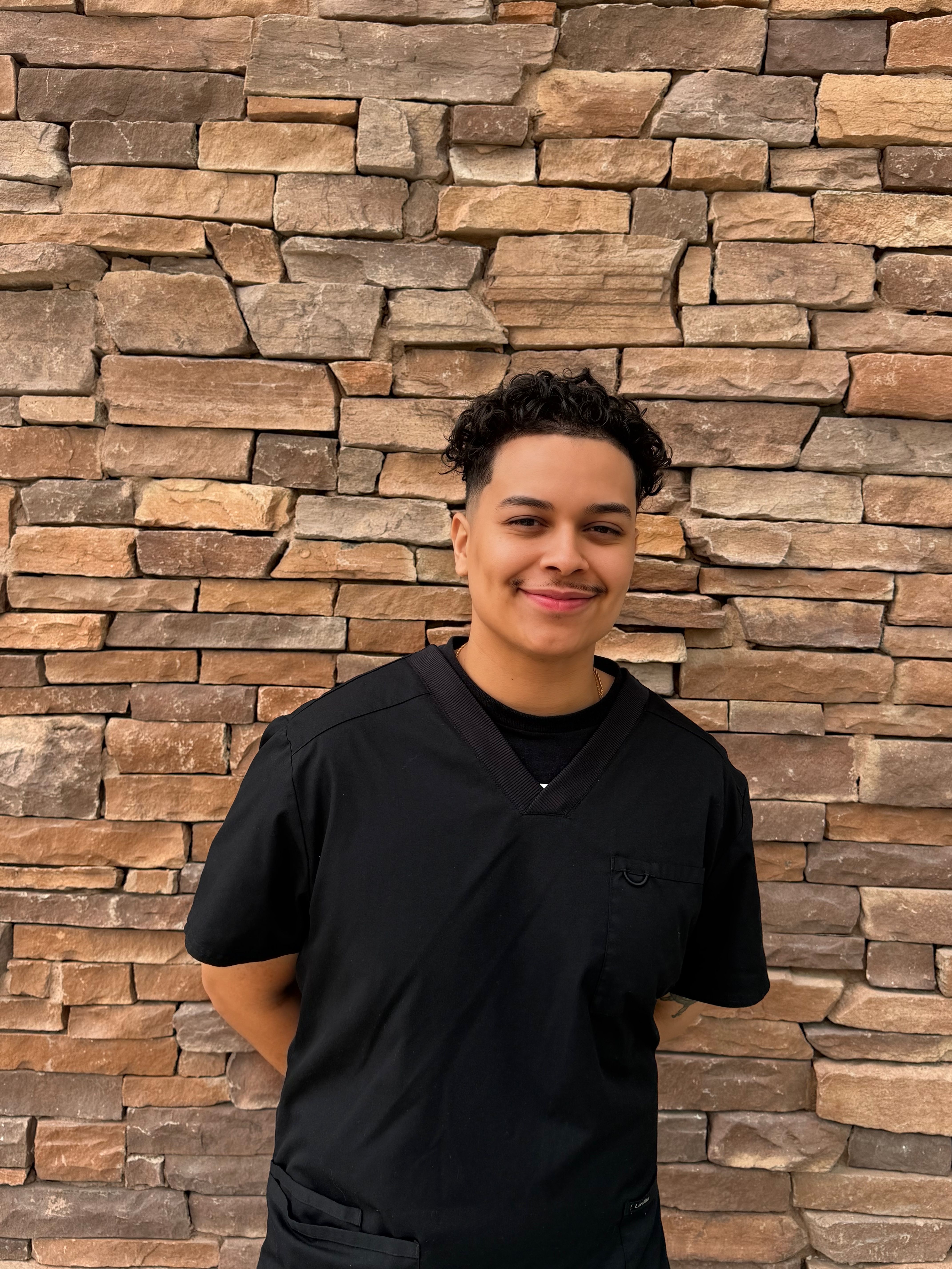 A smiling person in black scrubs stands against a textured stone wall.