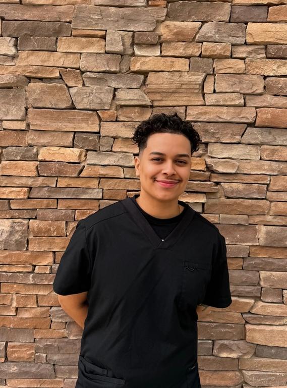 A smiling person in black scrubs stands against a textured stone wall.