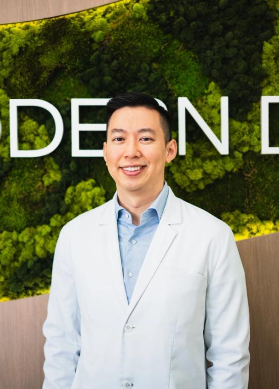 A smiling man in a white lab coat and blue shirt stands against a green moss wall with white letters spelling "MODERN".