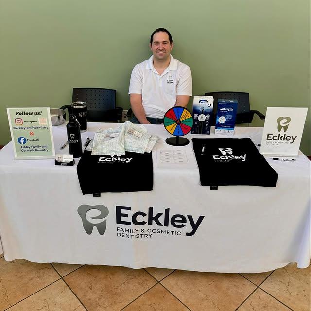 A man smiles behind a white table promoting Eckley Family & Cosmetic Dentistry, featuring logo shirts, a prize wheel, and dental product displays.