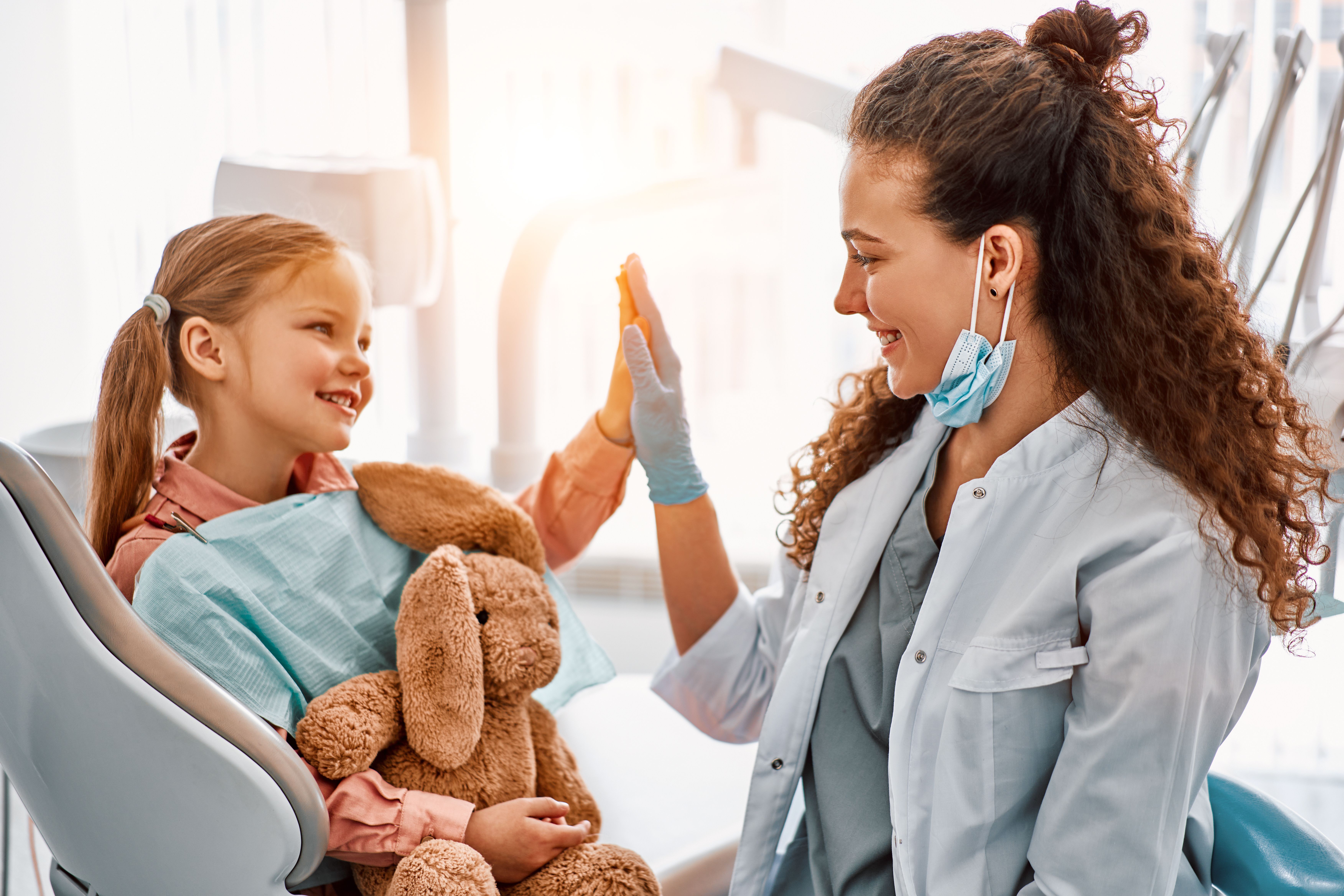 A cheerful young girl holding a stuffed bunny high-fives a smiling female healthcare worker.