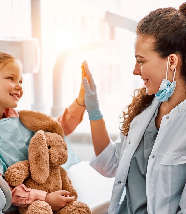 A cheerful young girl holding a stuffed bunny high-fives a smiling female healthcare worker.