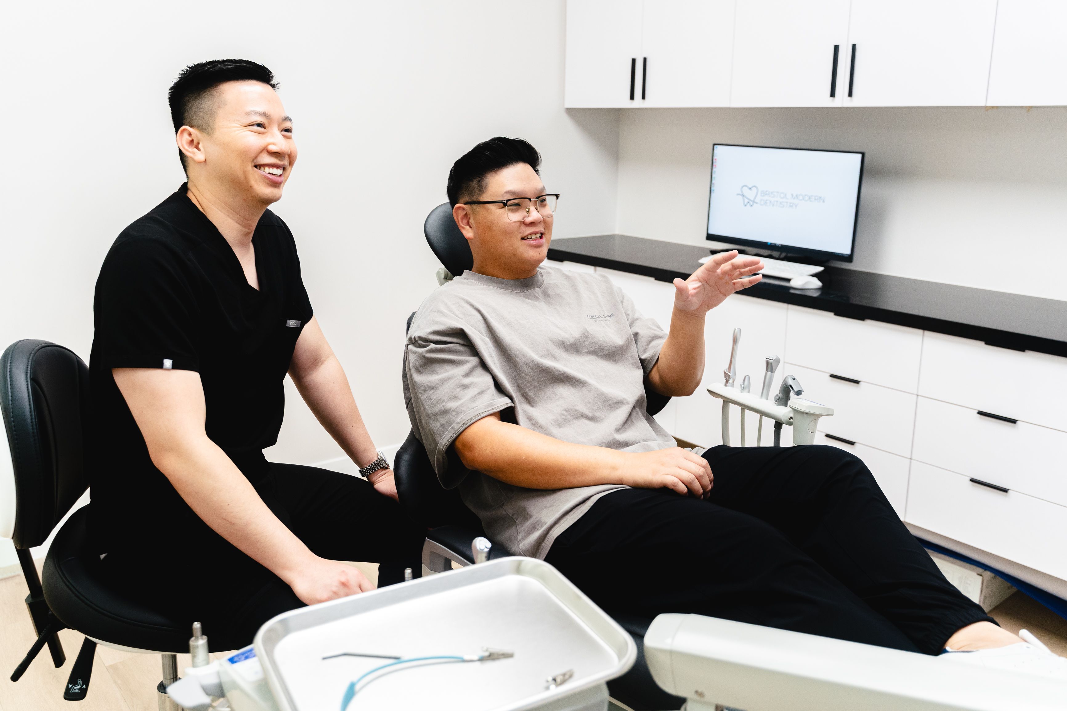 A smiling dentist in scrubs stands next to a patient in a dental chair who is talking and gesturing in a modern dental office.