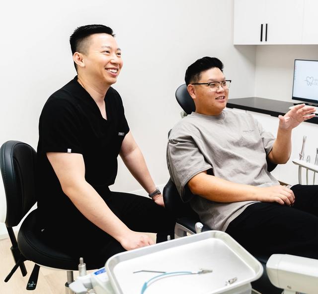A smiling dentist in scrubs stands next to a patient in a dental chair who is talking and gesturing in a modern dental office.
