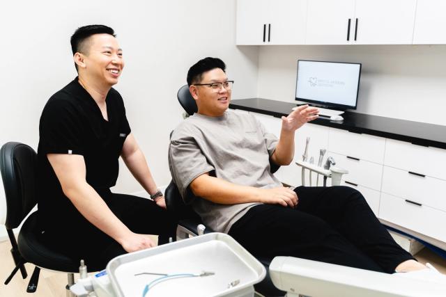 Two men, one in scrubs, smiling and conversing with a patient in a dental chair.