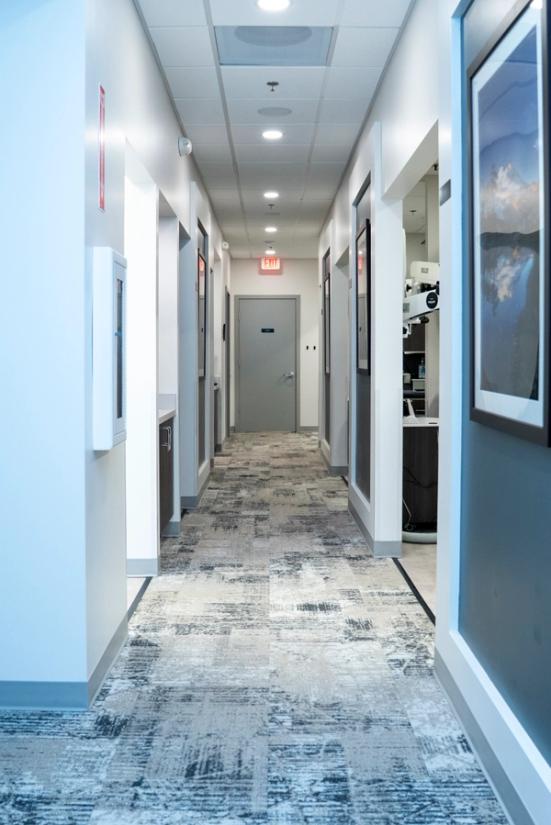 A brightly lit hallway with white walls, a gray patterned carpet, framed art, and an exit sign at the far end.