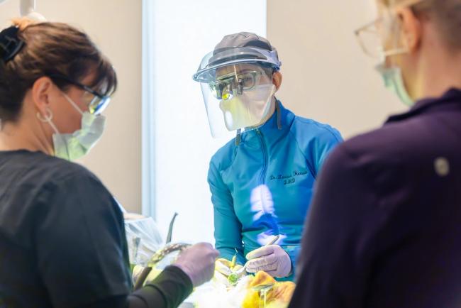 Three dental professionals, including a dentist in a face shield and loupes, performing a procedure.