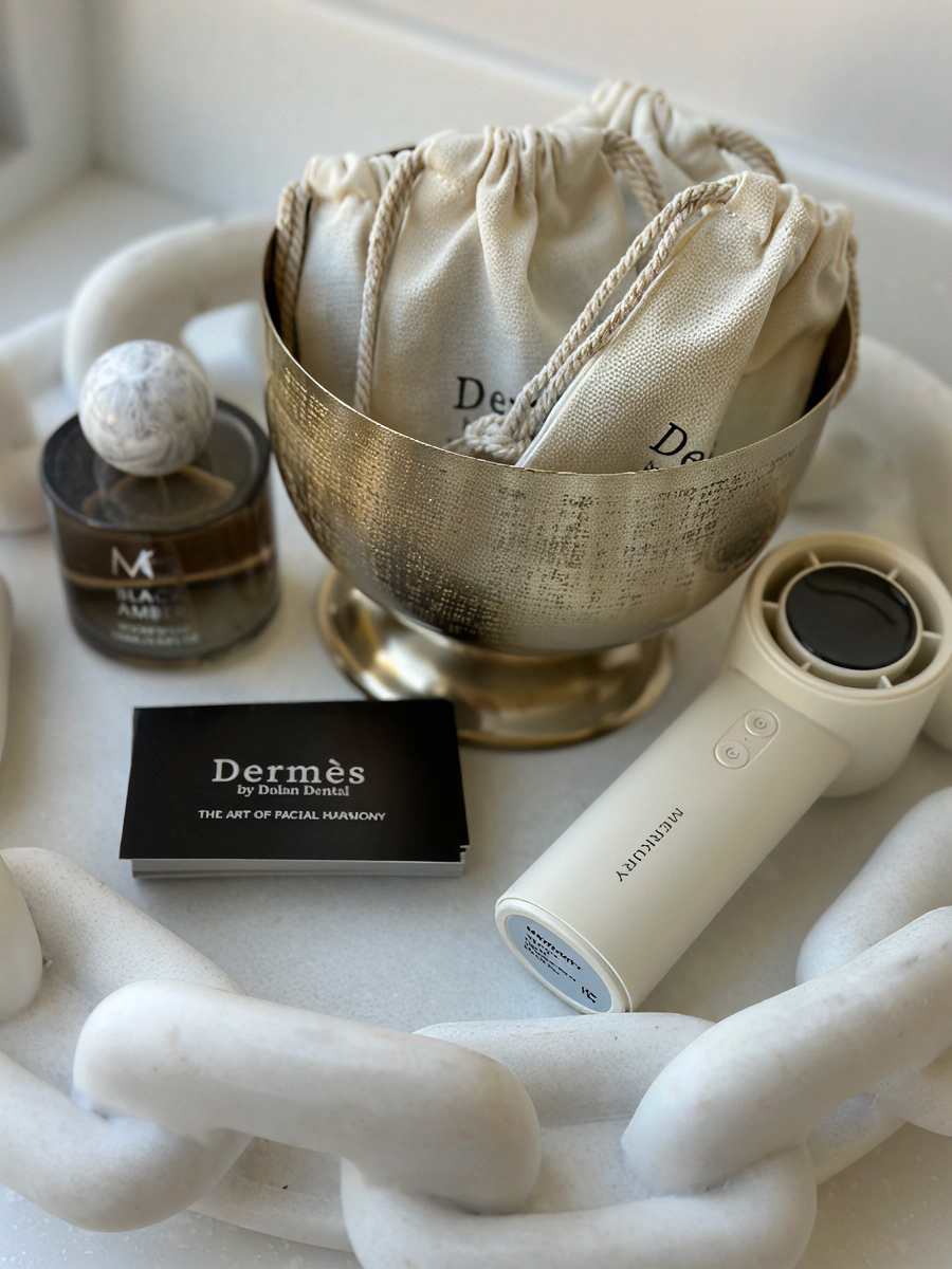 A brass bowl with "Dermès" pouches, a "Black Amber" perfume, a "Dermès" brochure, and a cream Merkury handheld device are arranged on a white decorative chain.