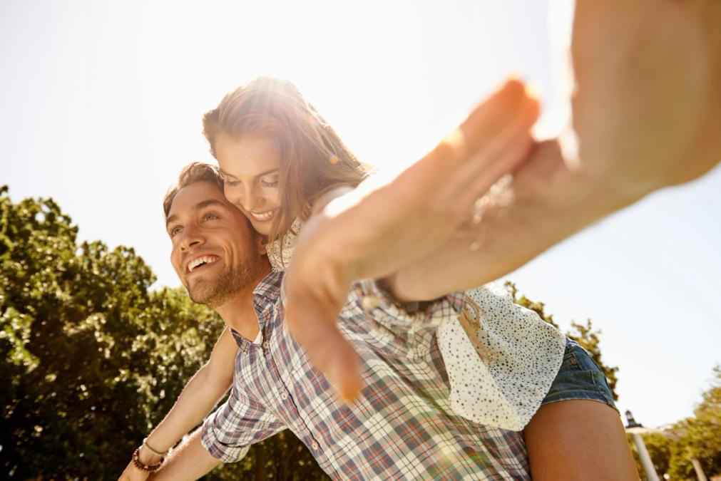 A happy couple on a sunny day, with the man giving the woman a piggyback ride as she reaches towards the camera.