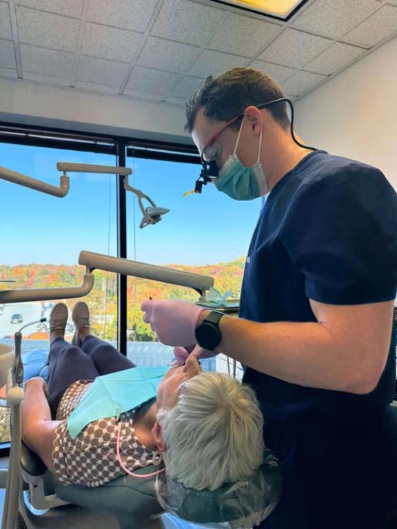 A dentist examines a patient in a dental chair, with a large window in the background showing fall foliage.