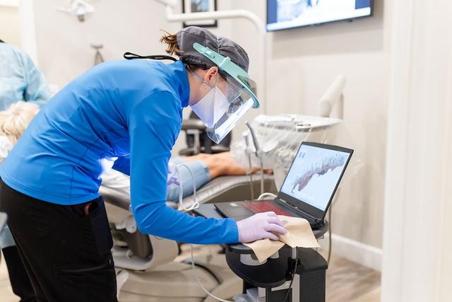 A dental professional in PPE uses a digital scanner connected to a laptop to create a 3D model of a patient's teeth.