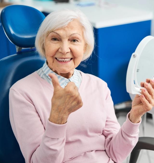 an elderly woman is sitting in a dental chair looking at her teeth in a mirror .