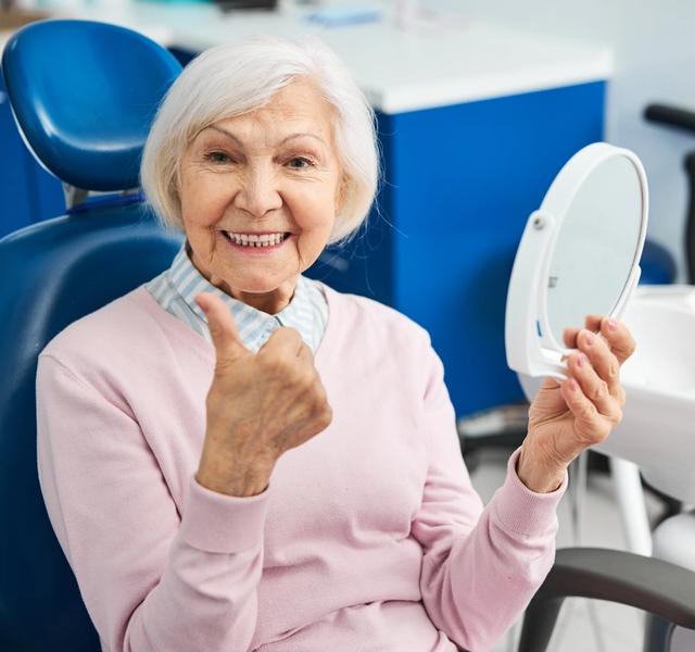 an elderly woman is sitting in a dental chair looking at her teeth in a mirror .