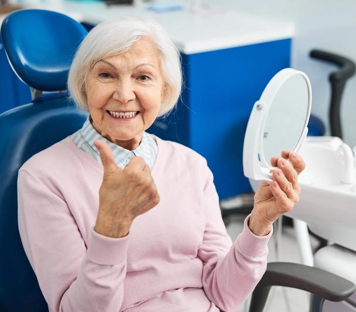 an elderly woman is sitting in a dental chair looking at her teeth in a mirror .