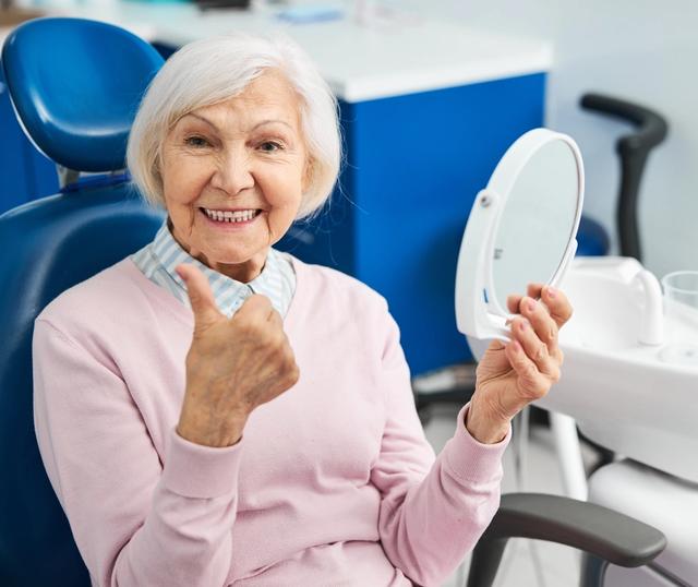 an elderly woman is sitting in a dental chair looking at her teeth in a mirror .