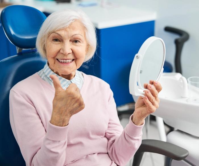 an elderly woman is sitting in a dental chair looking at her teeth in a mirror .