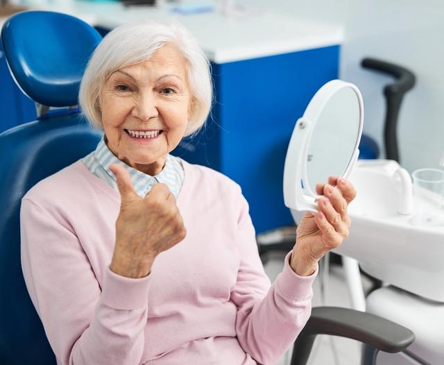 an elderly woman is sitting in a dental chair looking at her teeth in a mirror .