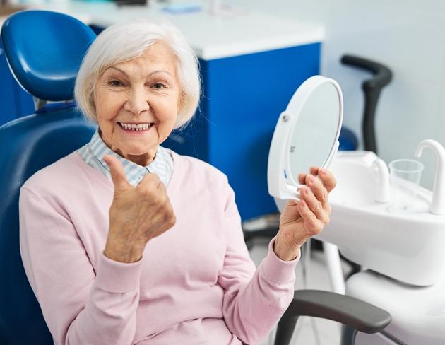 an elderly woman is sitting in a dental chair looking at her teeth in a mirror .