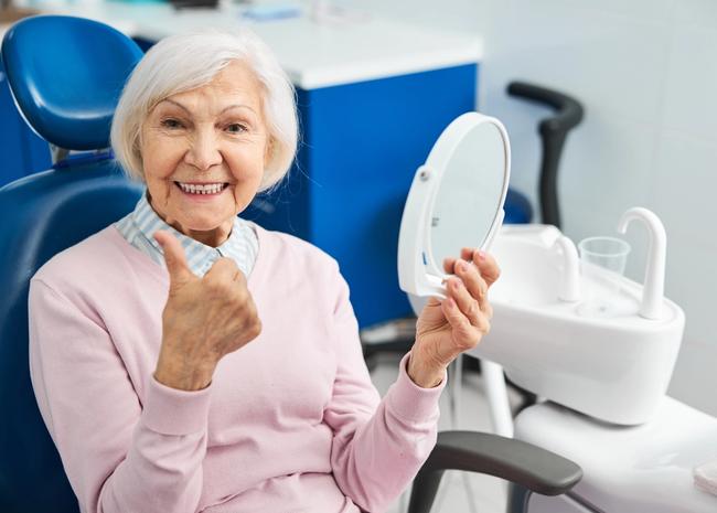 an elderly woman is sitting in a dental chair looking at her teeth in a mirror .