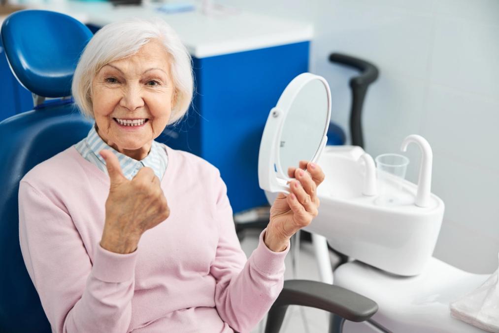 A smiling elderly woman in a dental chair gives a thumbs-up while holding a mirror.