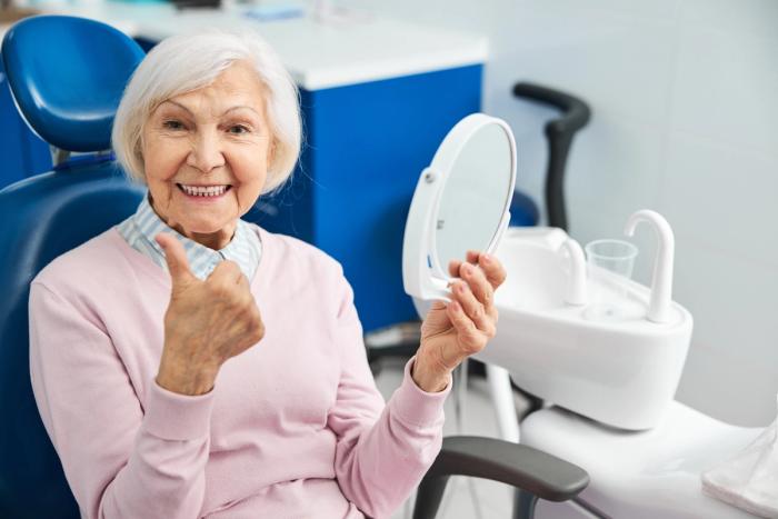 an elderly woman is sitting in a dental chair looking at her teeth in a mirror .