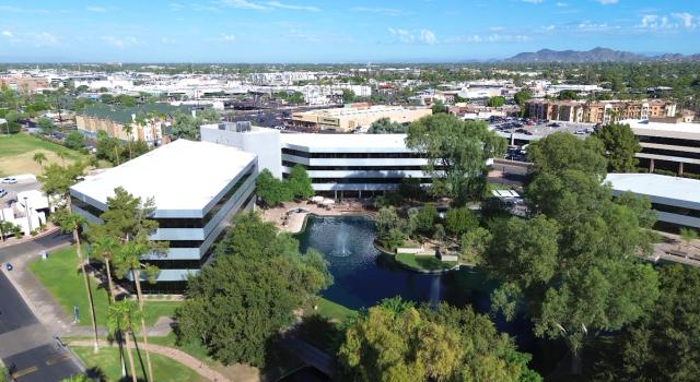 Aerial view of a business park featuring modern buildings, a pond with a fountain, lush trees, and a city with mountains in the background.