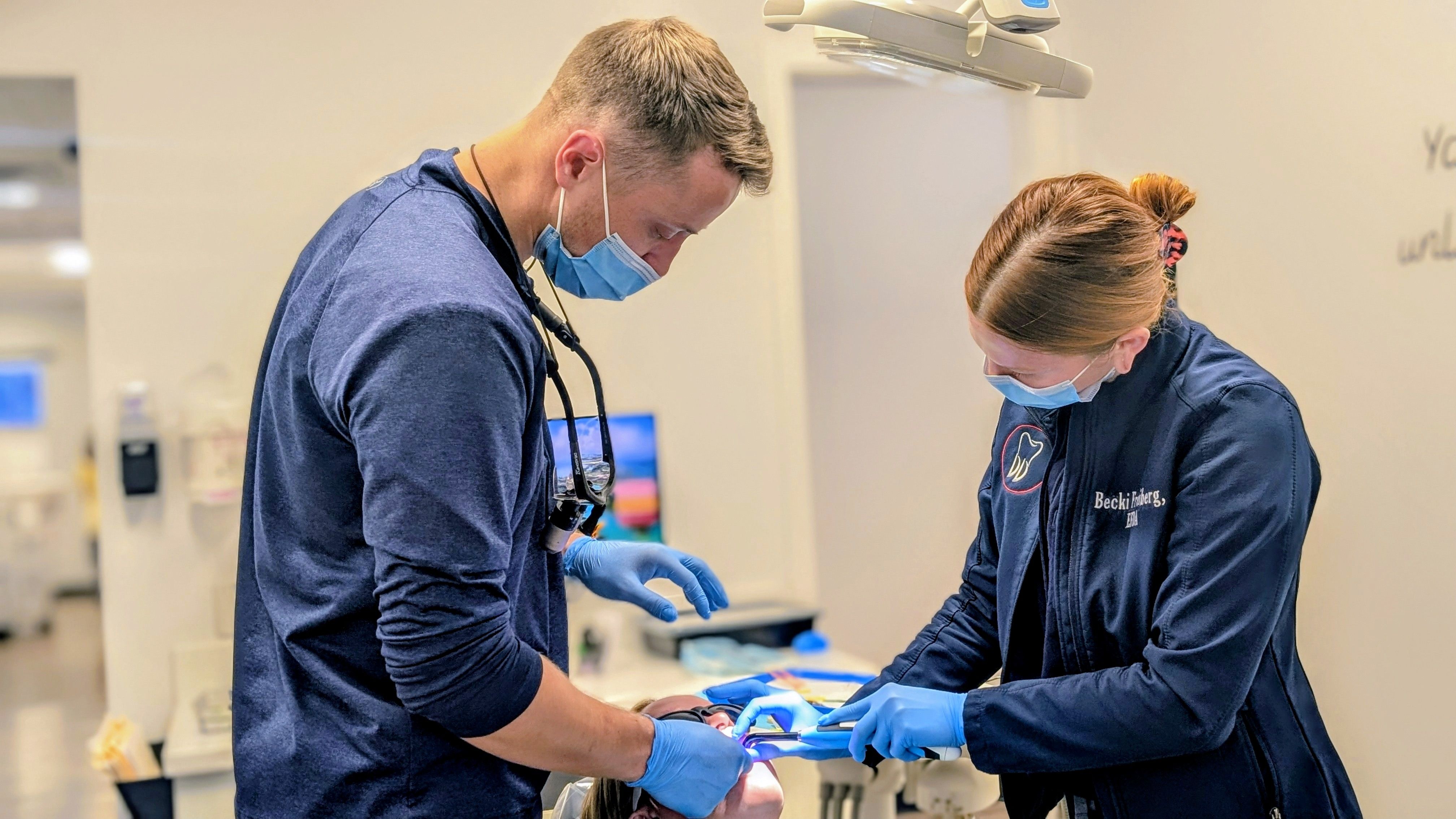 A male dentist and female assistant, both masked and gloved, performing a dental procedure on a patient.